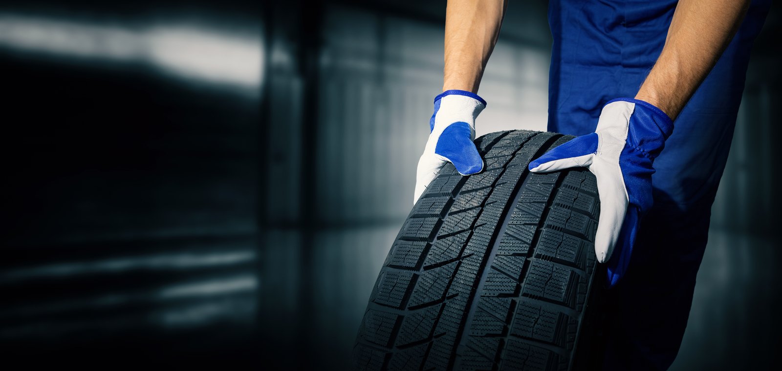 Car Tire Shop And Service Mechanic Holding New Tyre On Garage Background. Copy Space